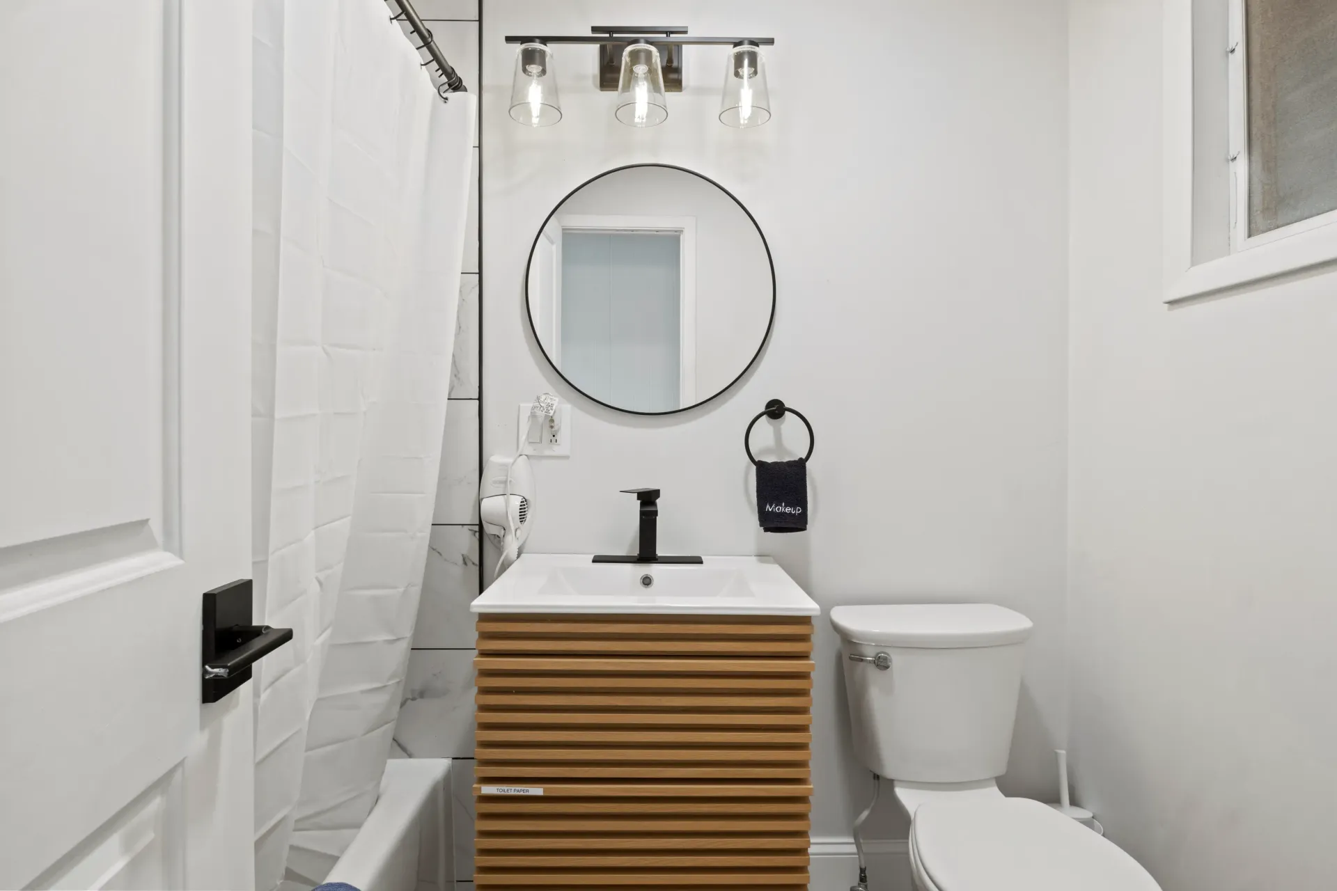 A modern bathroom featuring a wood vanity, round mirror, white toilet, and a bathtub with a white shower curtain.