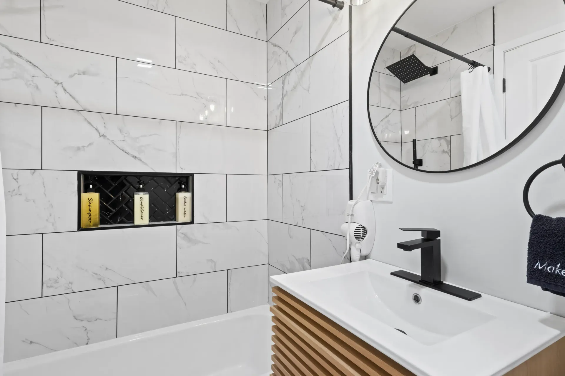 Modern bathroom with white marble-tiled walls, a slatted wood vanity, matte black fixtures, and a round wall mirror.