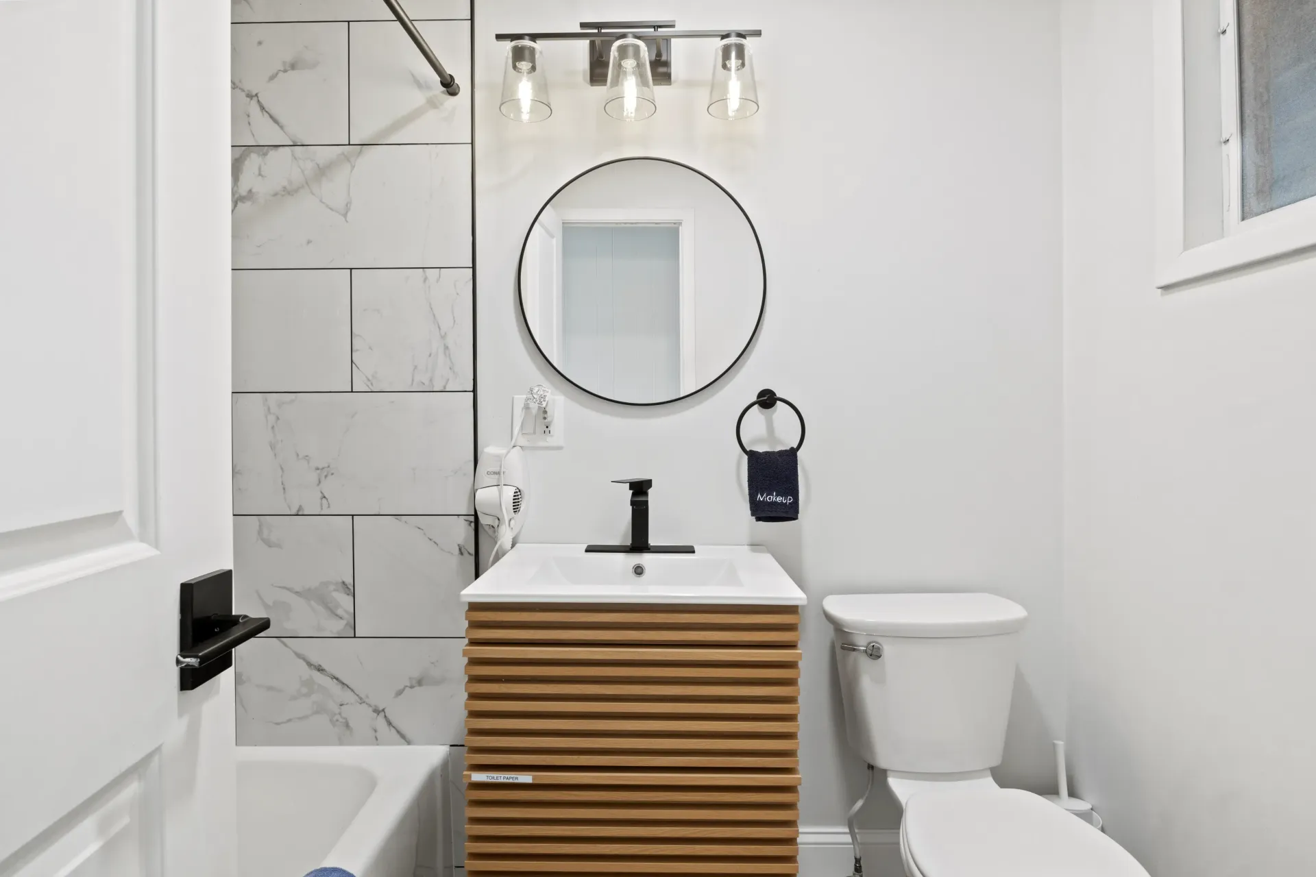 A small, modern bathroom featuring a white vanity with a wood-slatted front, round mirror, and a marbled tile shower area.
