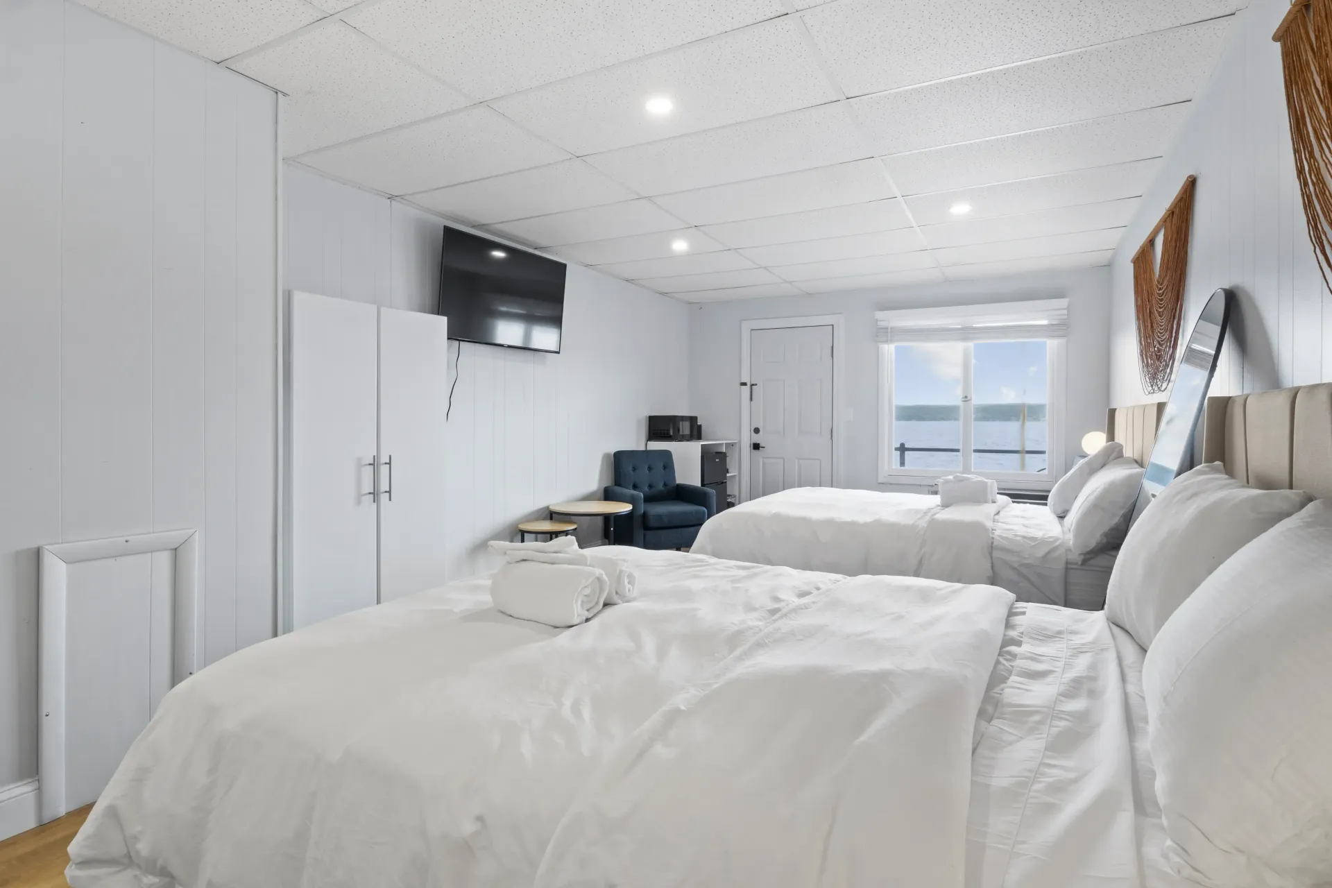 A hotel room featuring two beds with white bedding, a wall-mounted TV, a chair, a window with water views, and white decor.