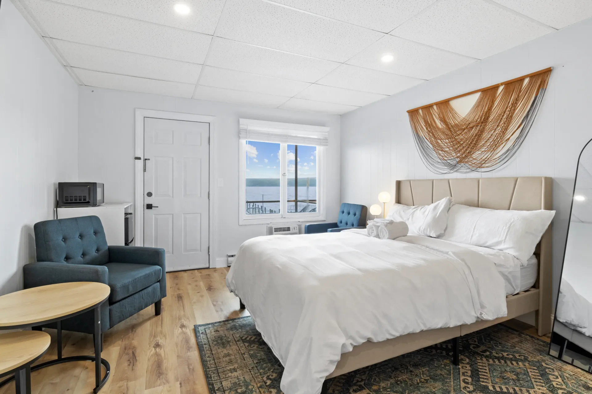 A brightly lit, neutral-toned bedroom with a queen bed, dark blue armchair, round side tables, and decorative wall art.