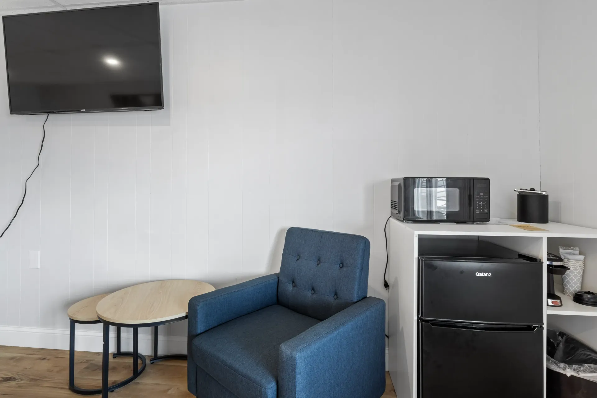 A room with a wall-mounted TV, a blue tufted armchair, a small side table, and a mini-fridge topped with a microwave.