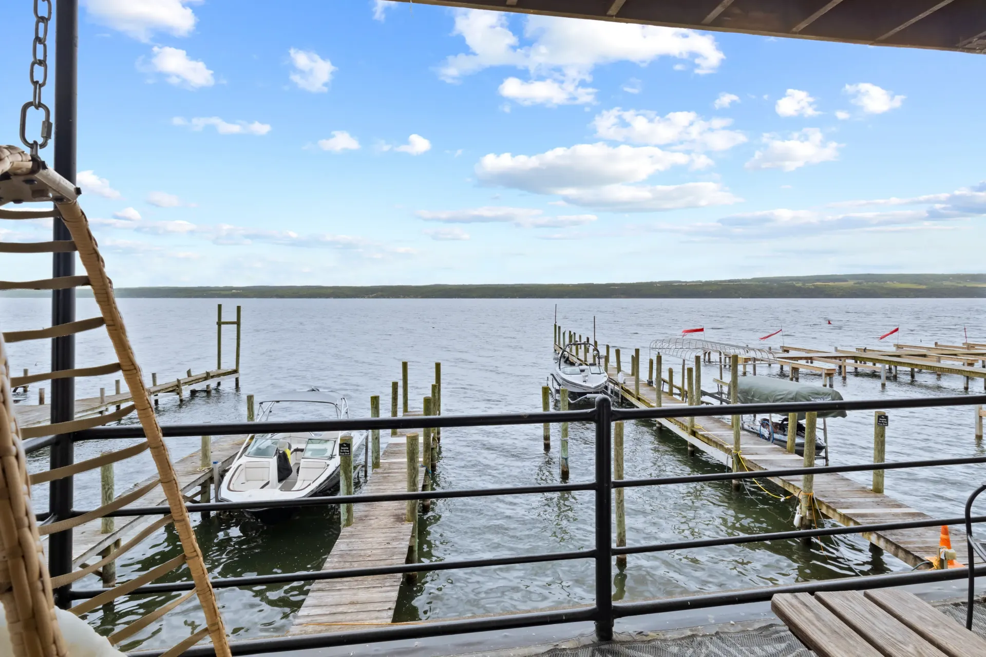 A view from a patio looking out at a lake with wooden docks, several boats, and a blue sky with scattered clouds.