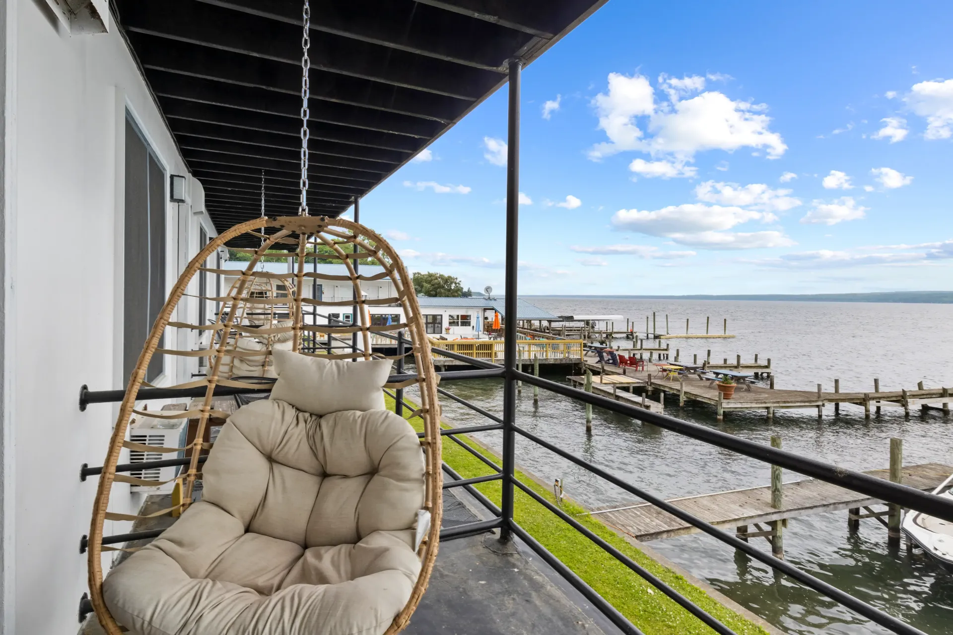 A beige wicker hanging chair on a balcony overlooking a lake with docks and a blue, partly cloudy sky.