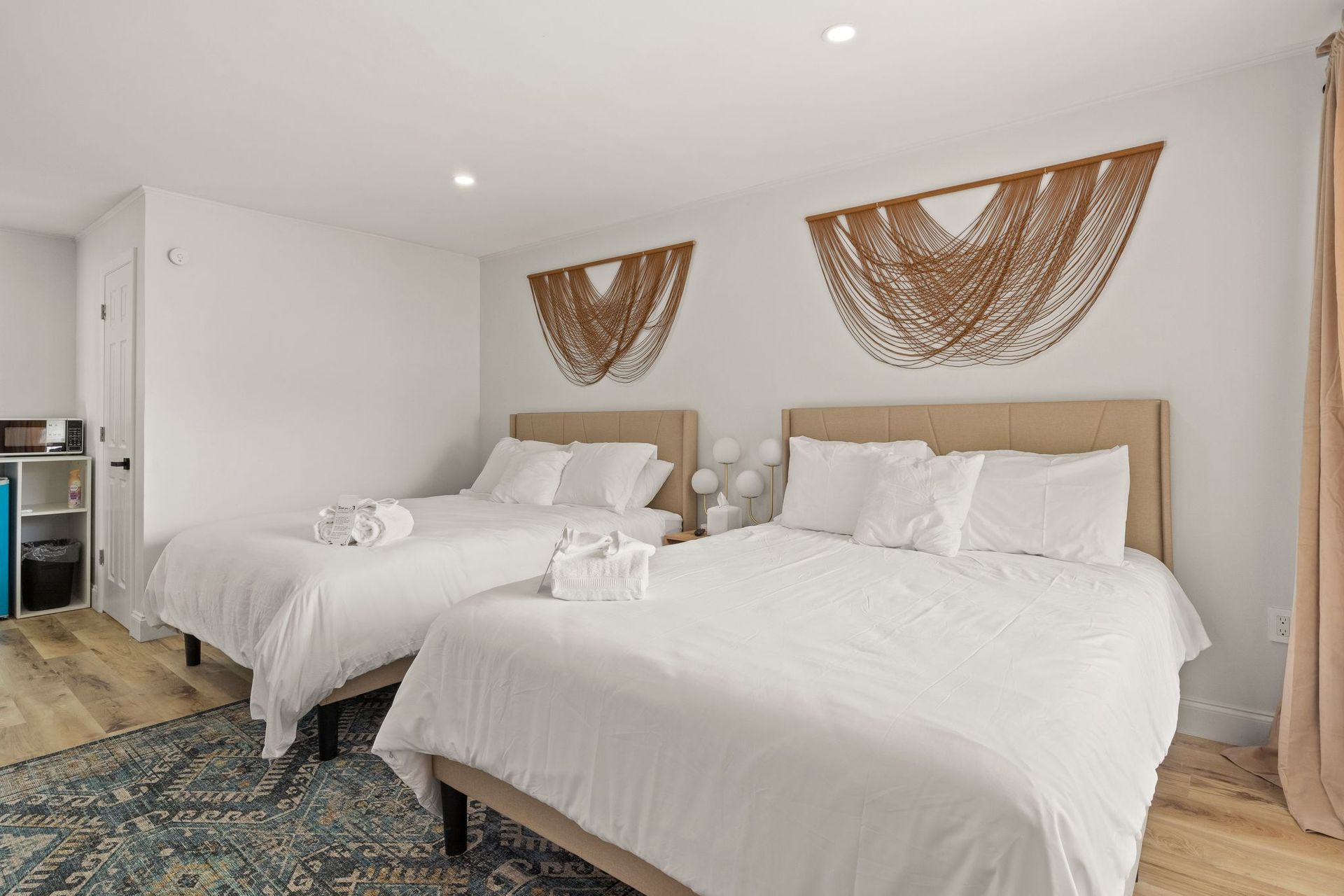 A brightly lit hotel room with two beds, white bedding, tan headboards, and decorative beaded wall hangings.