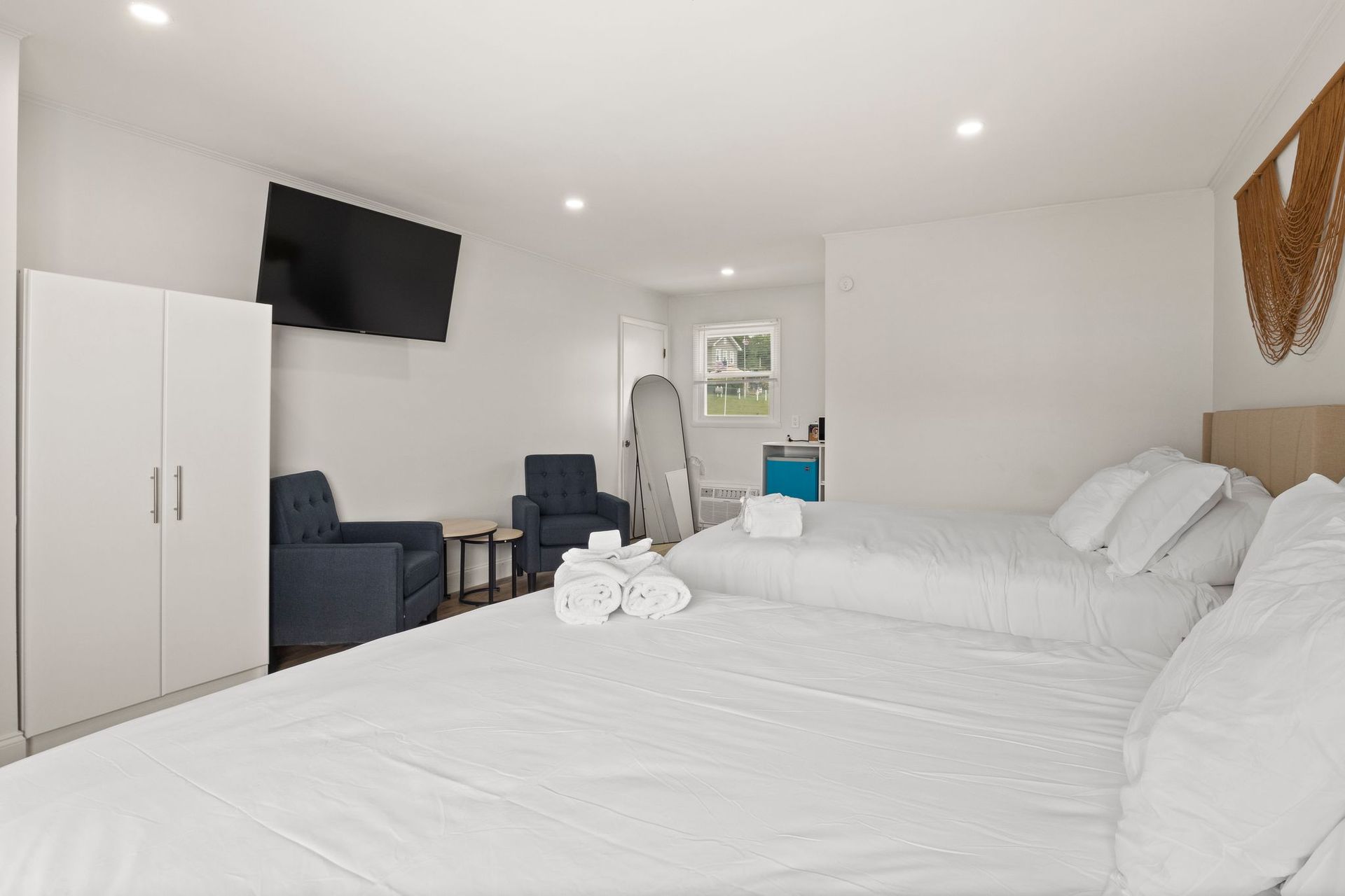 A hotel room with two white beds, a wardrobe, two blue armchairs, and a wall-mounted TV.