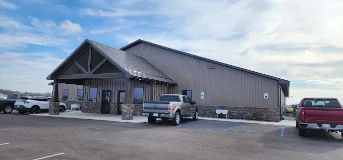 Small rustic building with parked trucks in a snowy parking lot under a cloudy sky