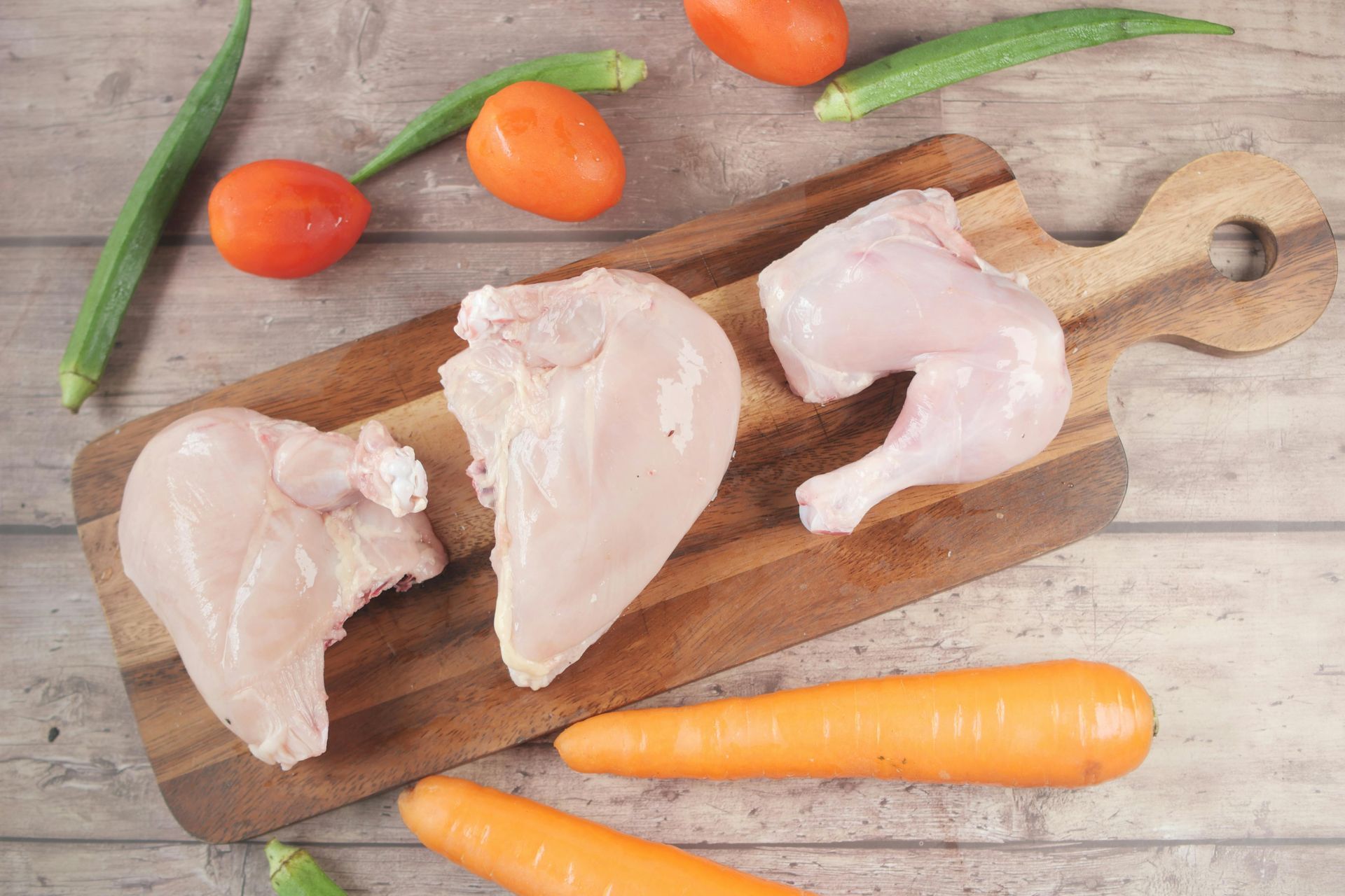 Raw chicken pieces on a wooden cutting board with carrots, tomatoes, and green onions on a rustic table