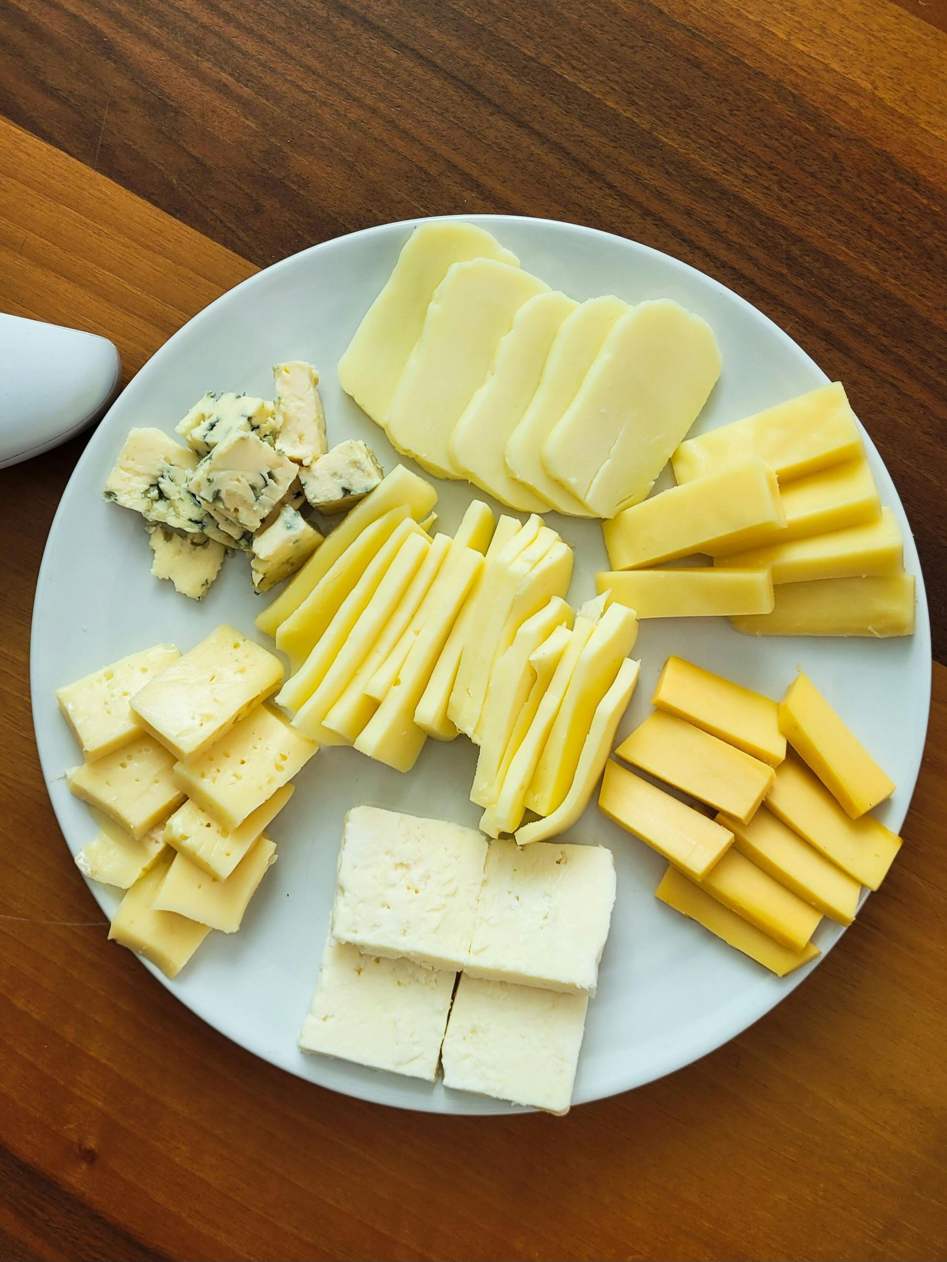 Assorted sliced cheeses arranged on a white plate on a wooden table.