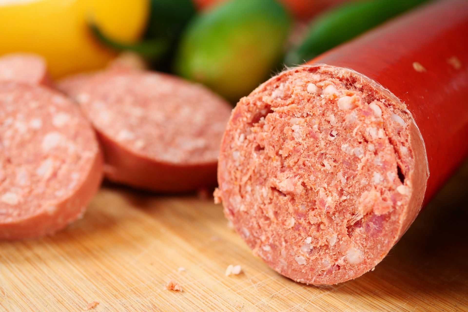 Sliced pink sausage log on a wooden cutting board with blurred fruit in the background