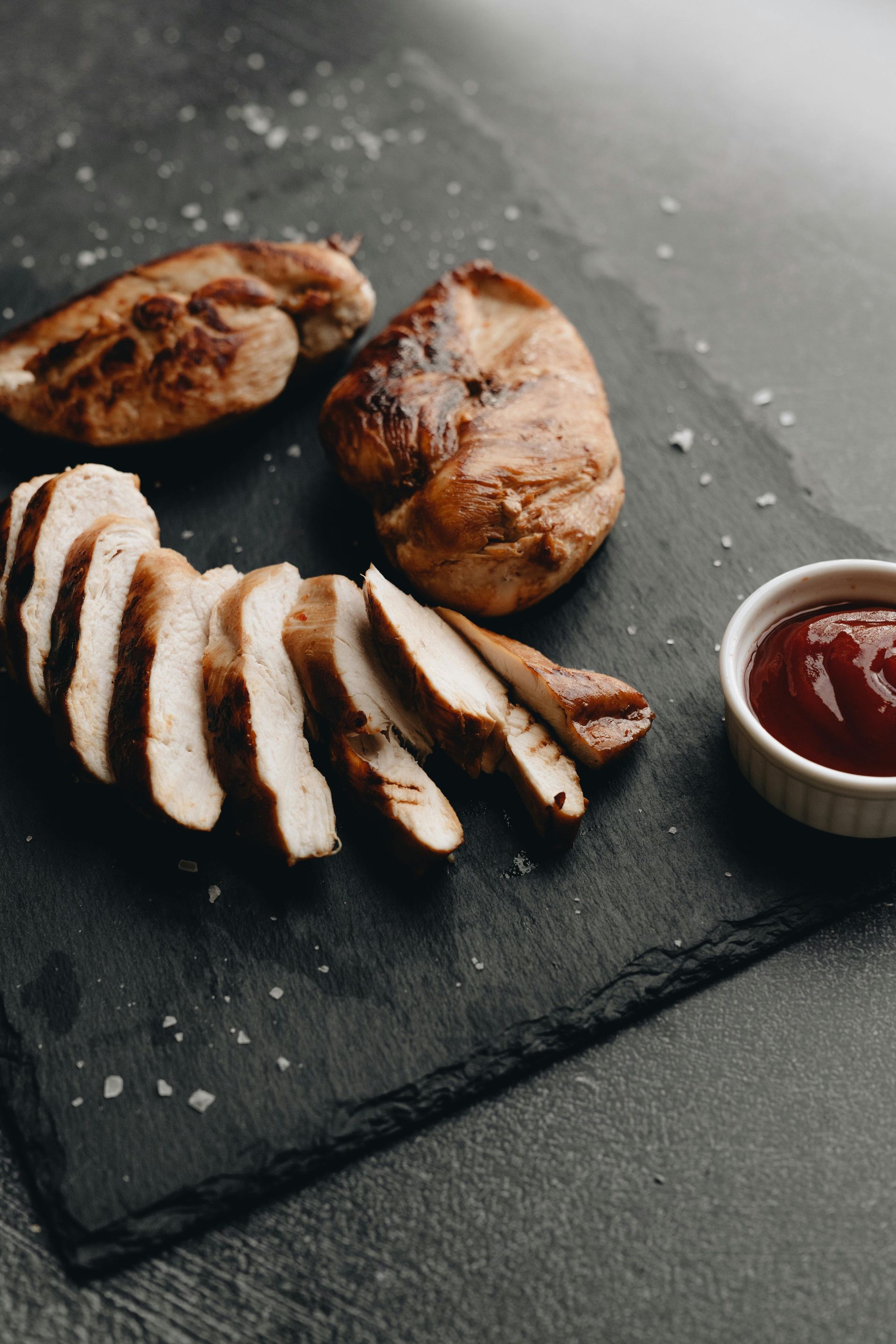 Sliced grilled chicken breast on a dark slate board with a small bowl of red sauce