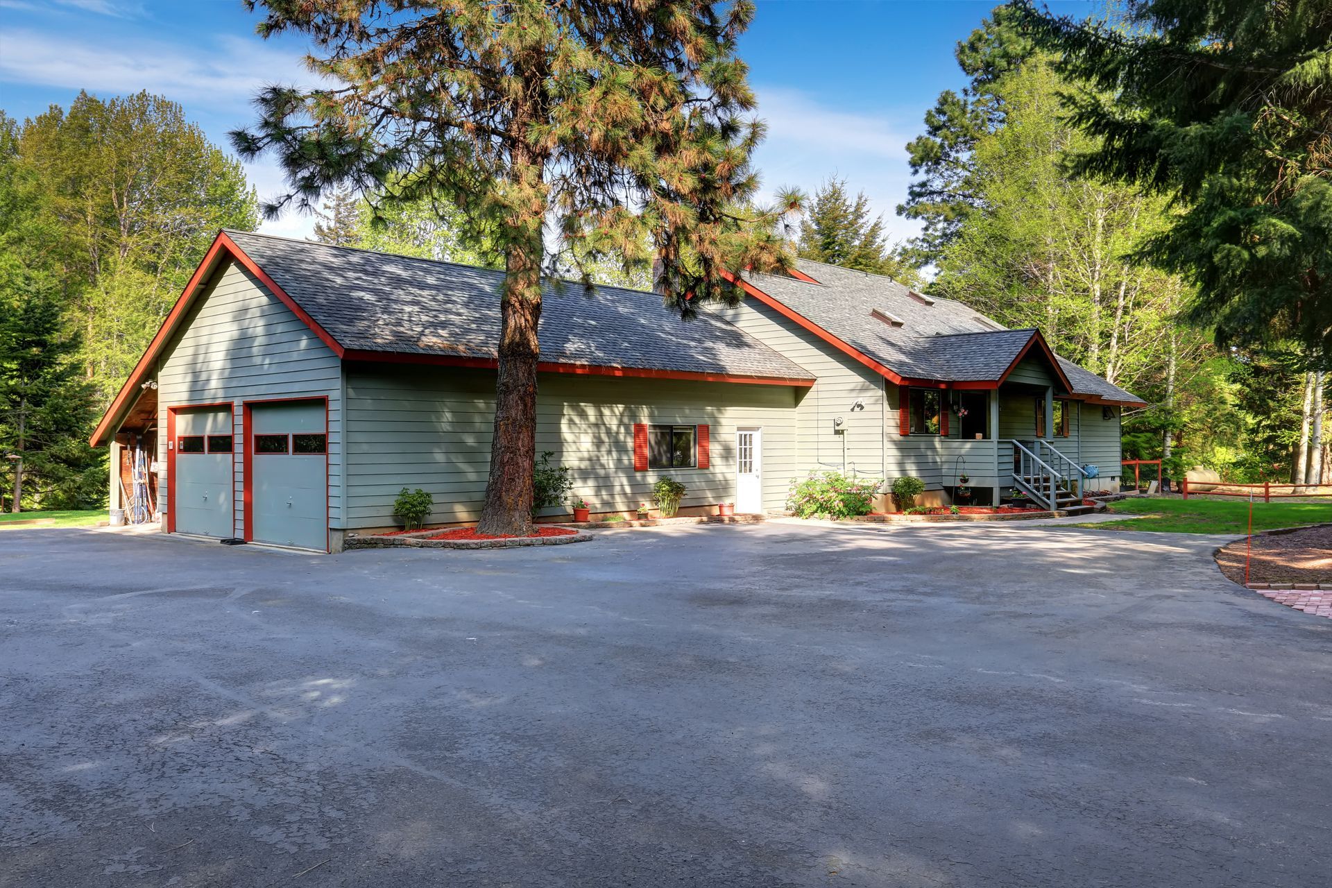 Gray house with red trim and garage doors amid trees, viewed from a gravel driveway Gray house with red trim and garage doors amid trees, viewed from a gravel driveway
