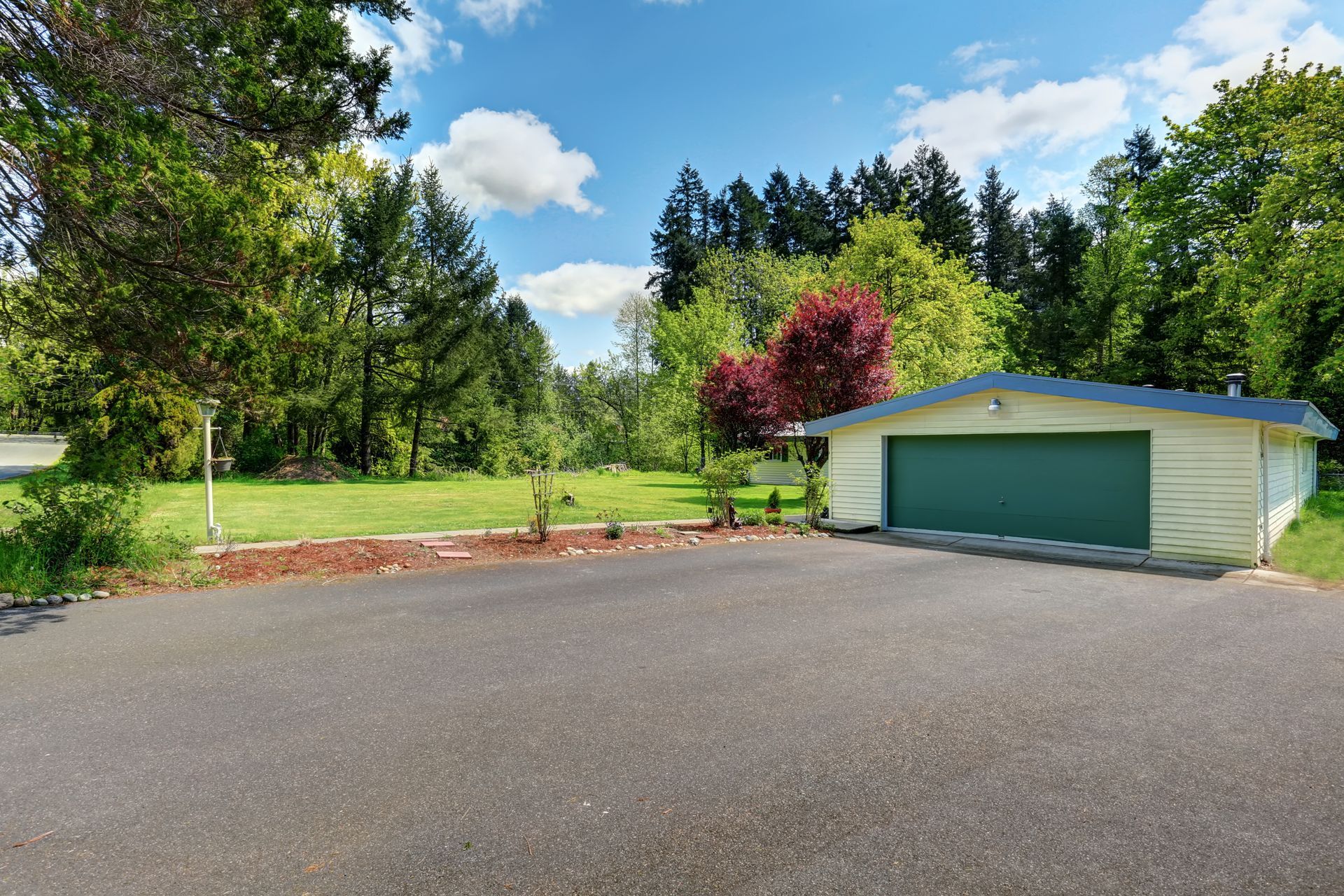 Driveway leading to a green garage beside a landscaped yard with trees and a blue sky. Driveway leading to a green garage beside a landscaped yard with trees and a blue sky.