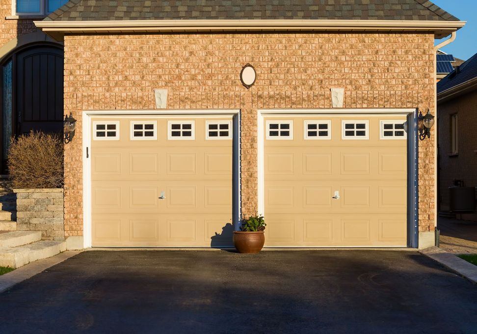 Two beige garage doors on a brick house with a potted plant between them Two beige garage doors on a brick house with a potted plant between them
