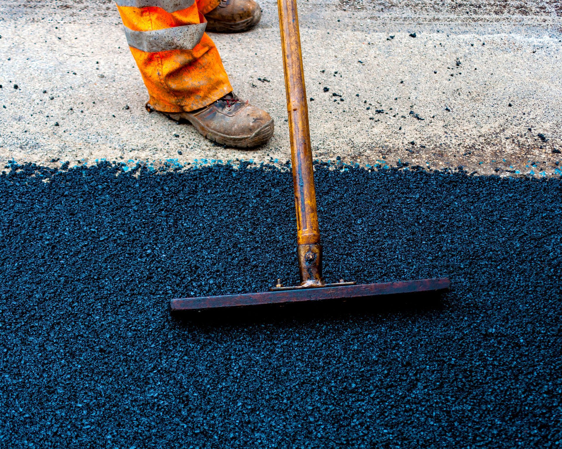Worker smoothing black asphalt with a rake near orange safety pants on a road surface Worker smoothing black asphalt with a rake near orange safety pants on a road surface
