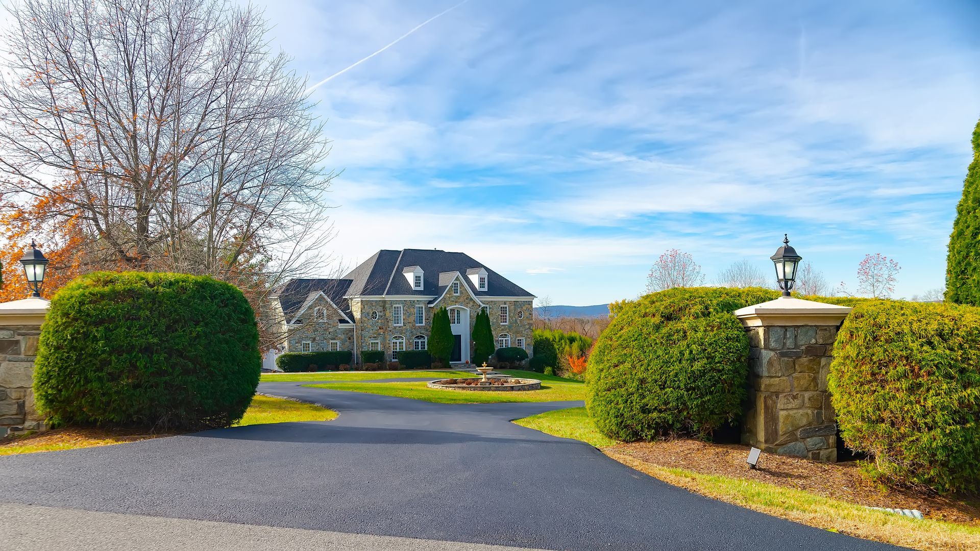 Estate driveway with manicured hedges, stone gateposts, and a large house under a blue sky Estate driveway with manicured hedges, stone gateposts, and a large house under a blue sky
