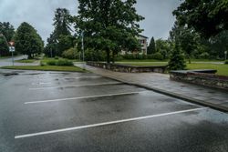 Rainy empty parking lot with wet asphalt, trees, and a small stone wall beside a grassy park Rainy empty parking lot with wet asphalt, trees, and a small stone wall beside a grassy park