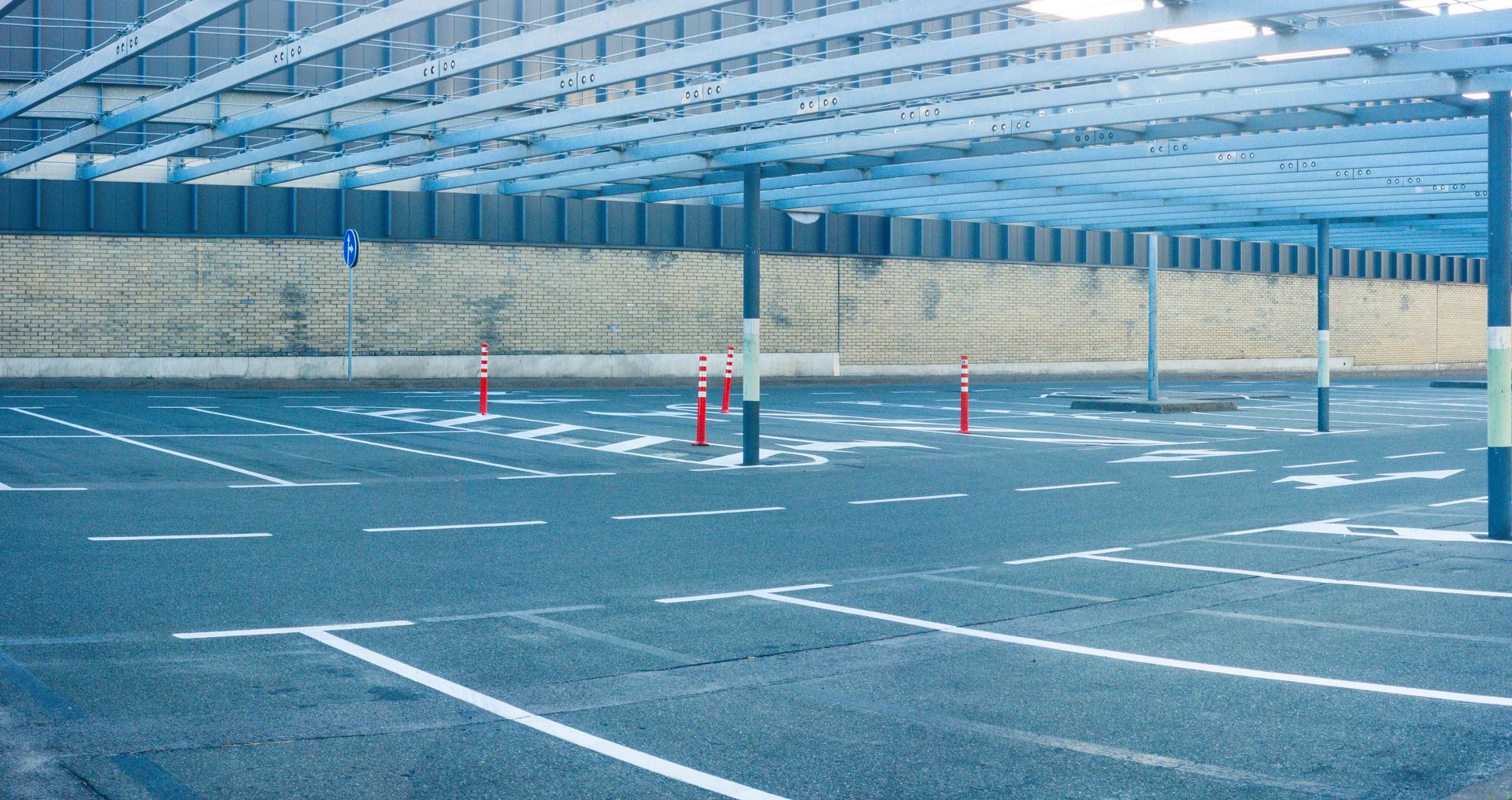 Empty covered parking lot with blue-painted asphalt, white lines, and orange cones under a metal roof Empty covered parking lot with blue-painted asphalt, white lines, and orange cones under a metal roof