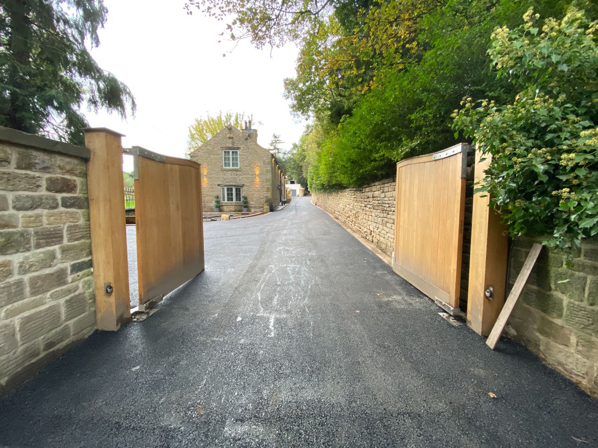 Stone driveway with open wooden gates leading to a house, lined by trees and stone walls. Stone driveway with open wooden gates leading to a house, lined by trees and stone walls.