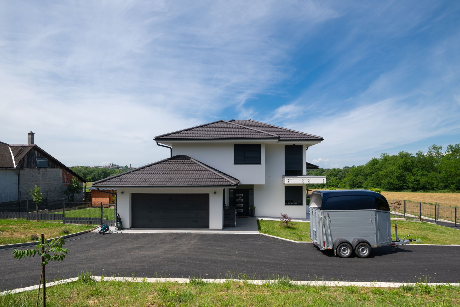 Modern two-story house with gray roof and a trailer parked in the driveway under a blue sky Modern two-story house with gray roof and a trailer parked in the driveway under a blue sky