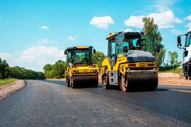 Two yellow road rollers paving a new asphalt road under a blue sky