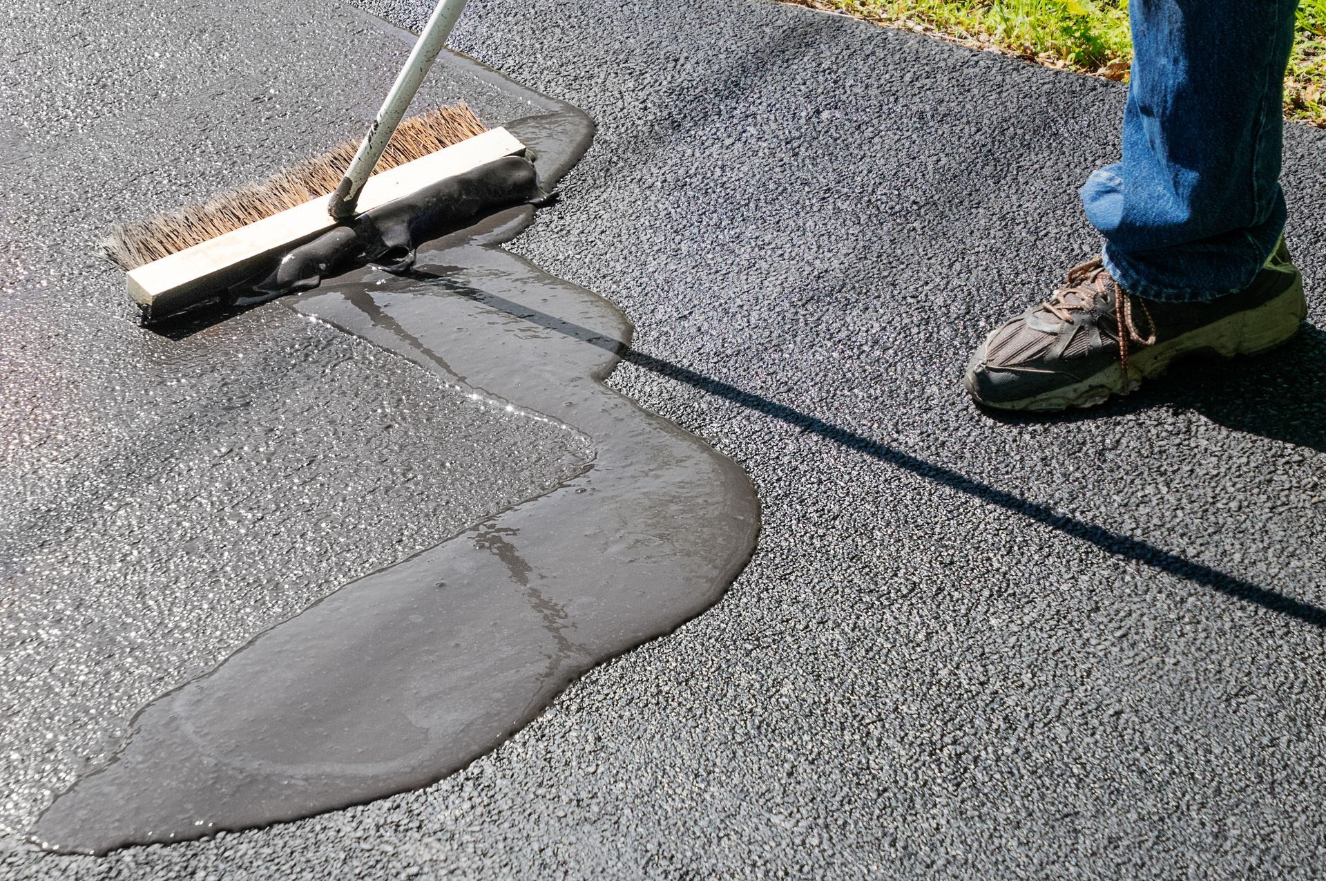 Person smoothing wet black sealant on pavement with a squeegee, next to a shoe-clad foot.