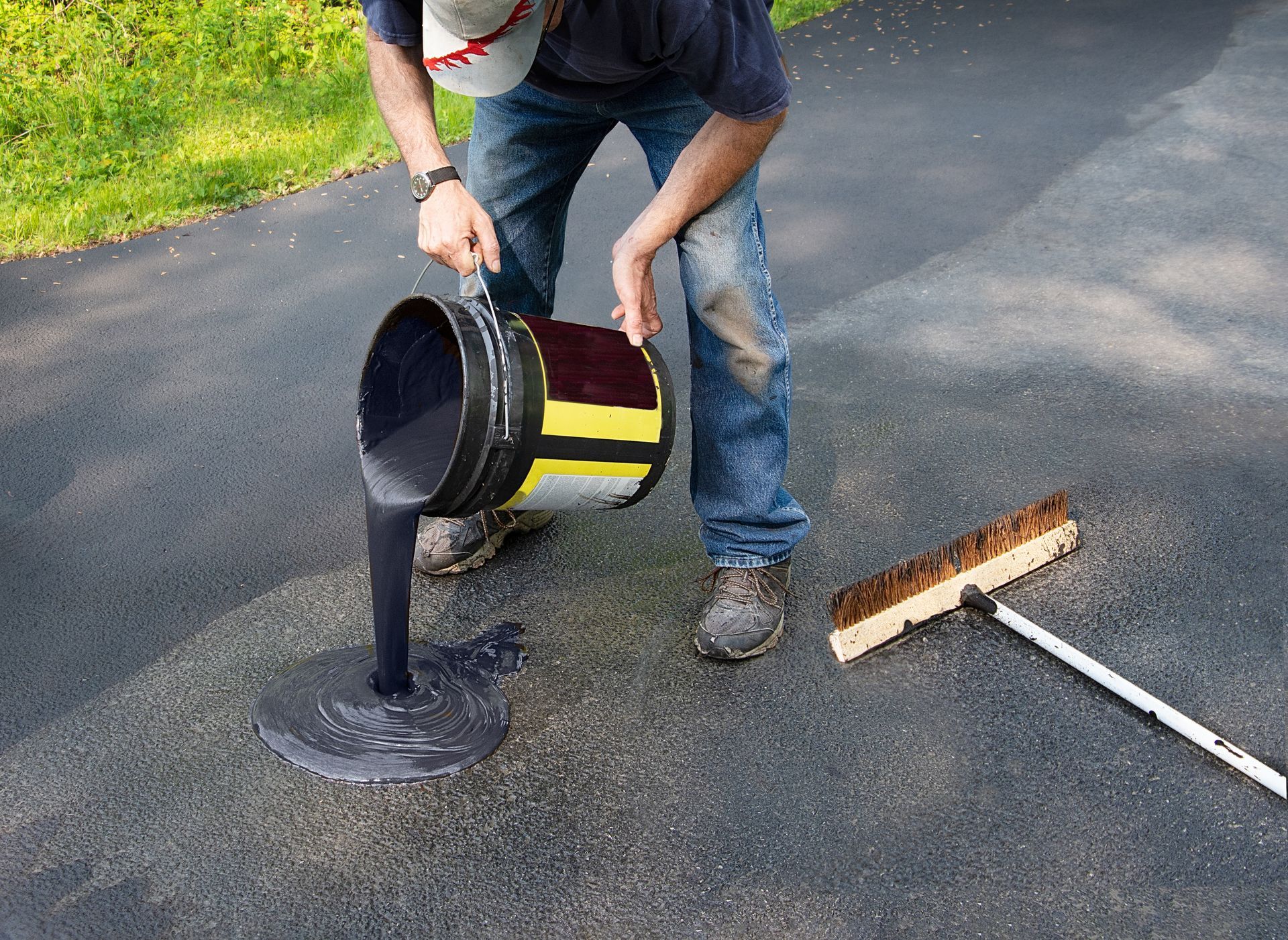 Person pouring black liquid from a yellow bucket onto a driveway beside a broom and shovel. Person pouring black liquid from a yellow bucket onto a driveway beside a broom and shovel.
