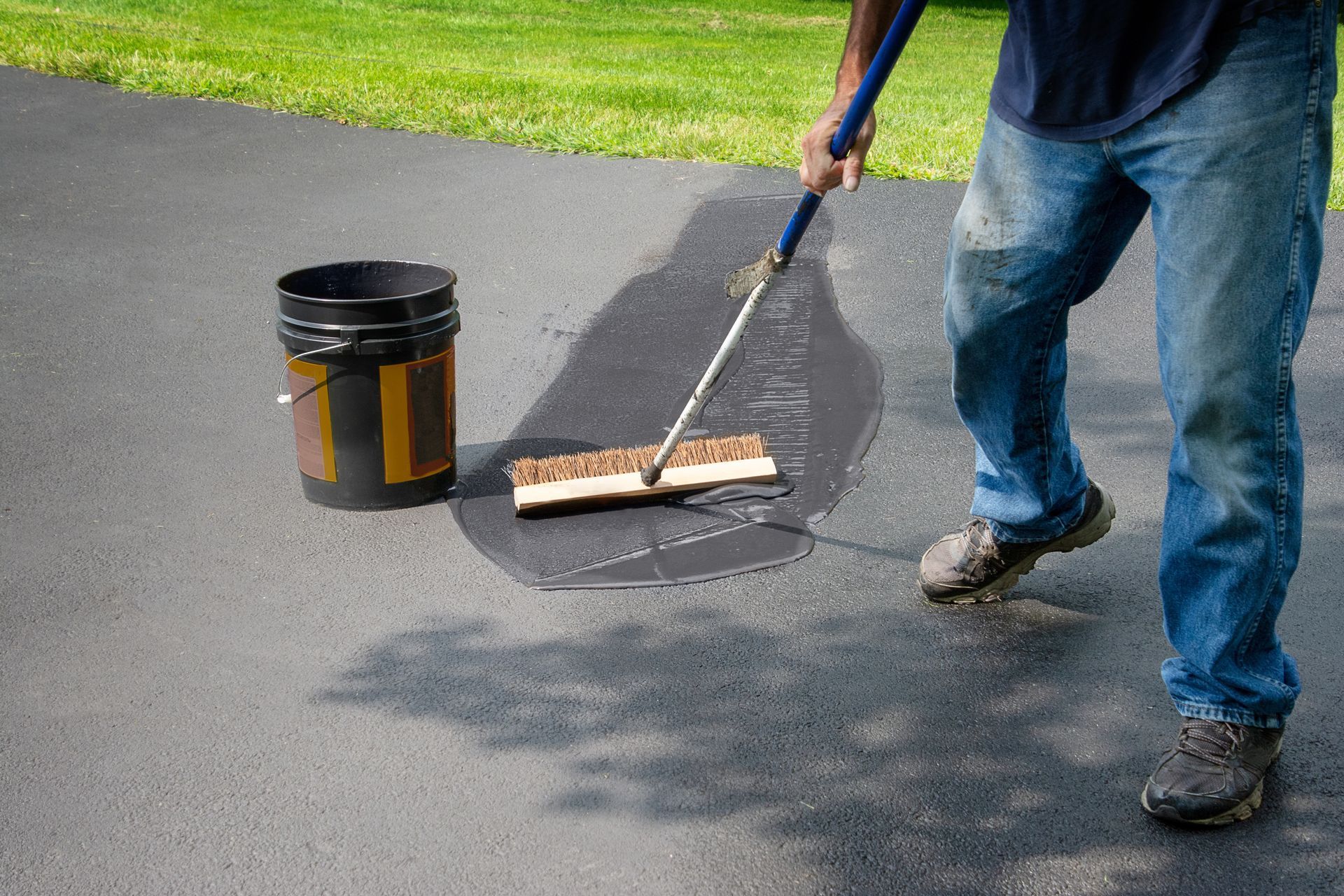 Person painting a crack on asphalt pavement with a roller beside a black paint bucket