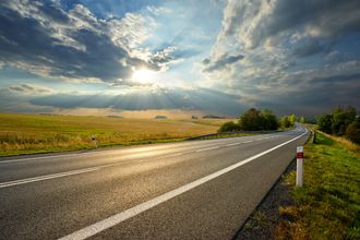 Country road at sunset under dramatic clouds, with golden fields and a reflective wet pavement