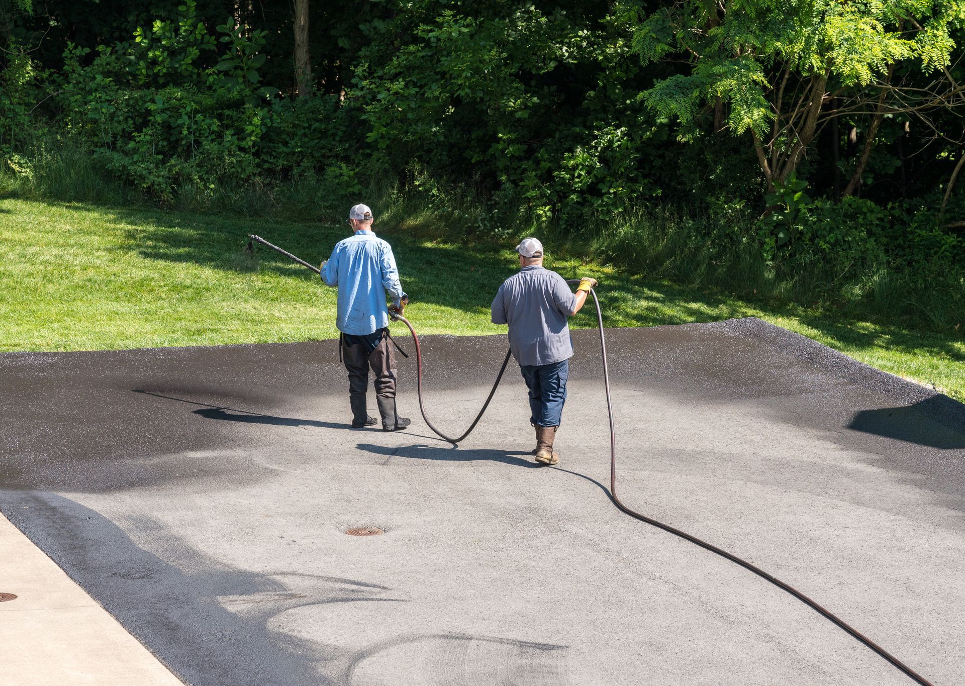 Two workers seal cracks in an asphalt road beside a grassy embankment. Two workers seal cracks in an asphalt road beside a grassy embankment.