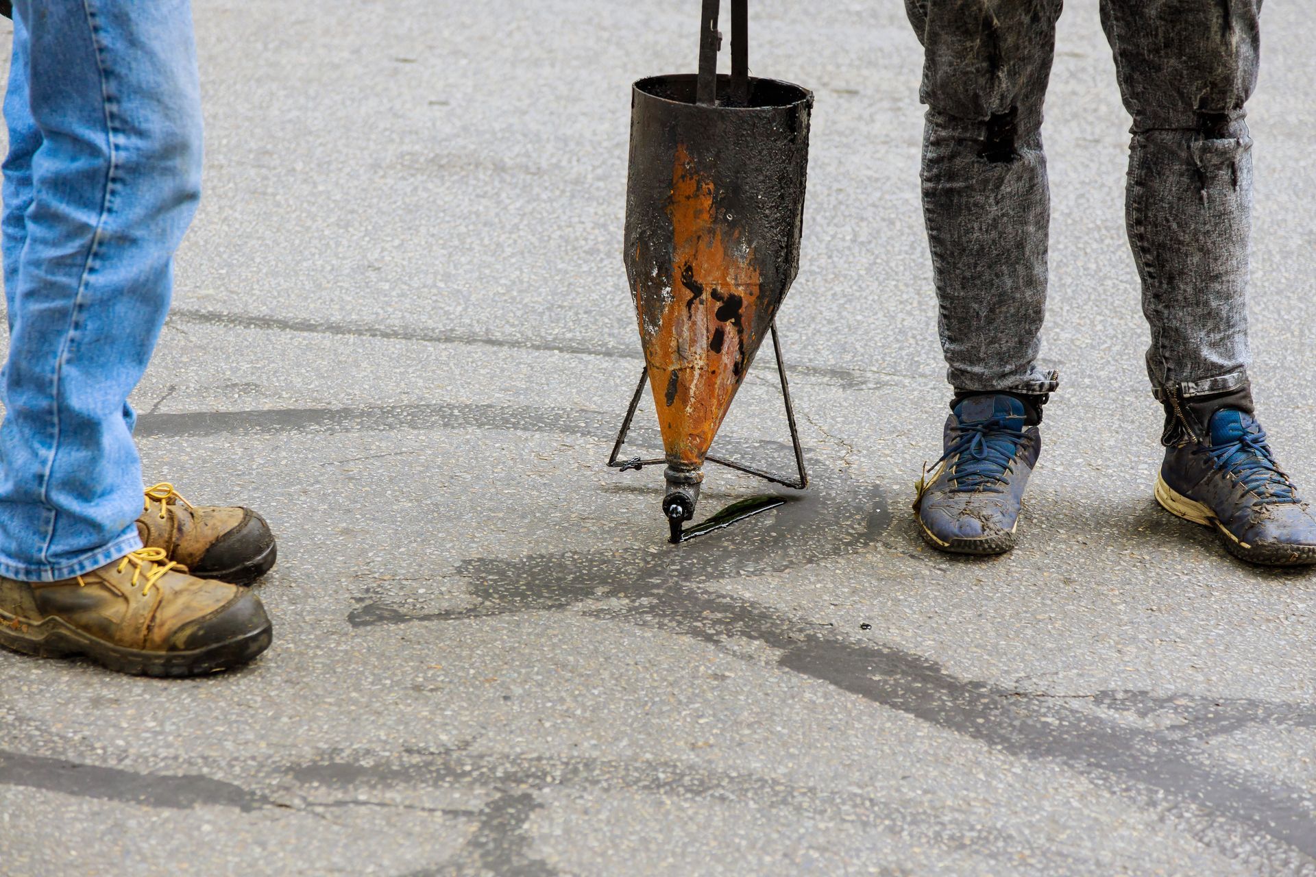 Workers standing around a pavement crack repair tool on wet asphalt, with shovels and boots visible