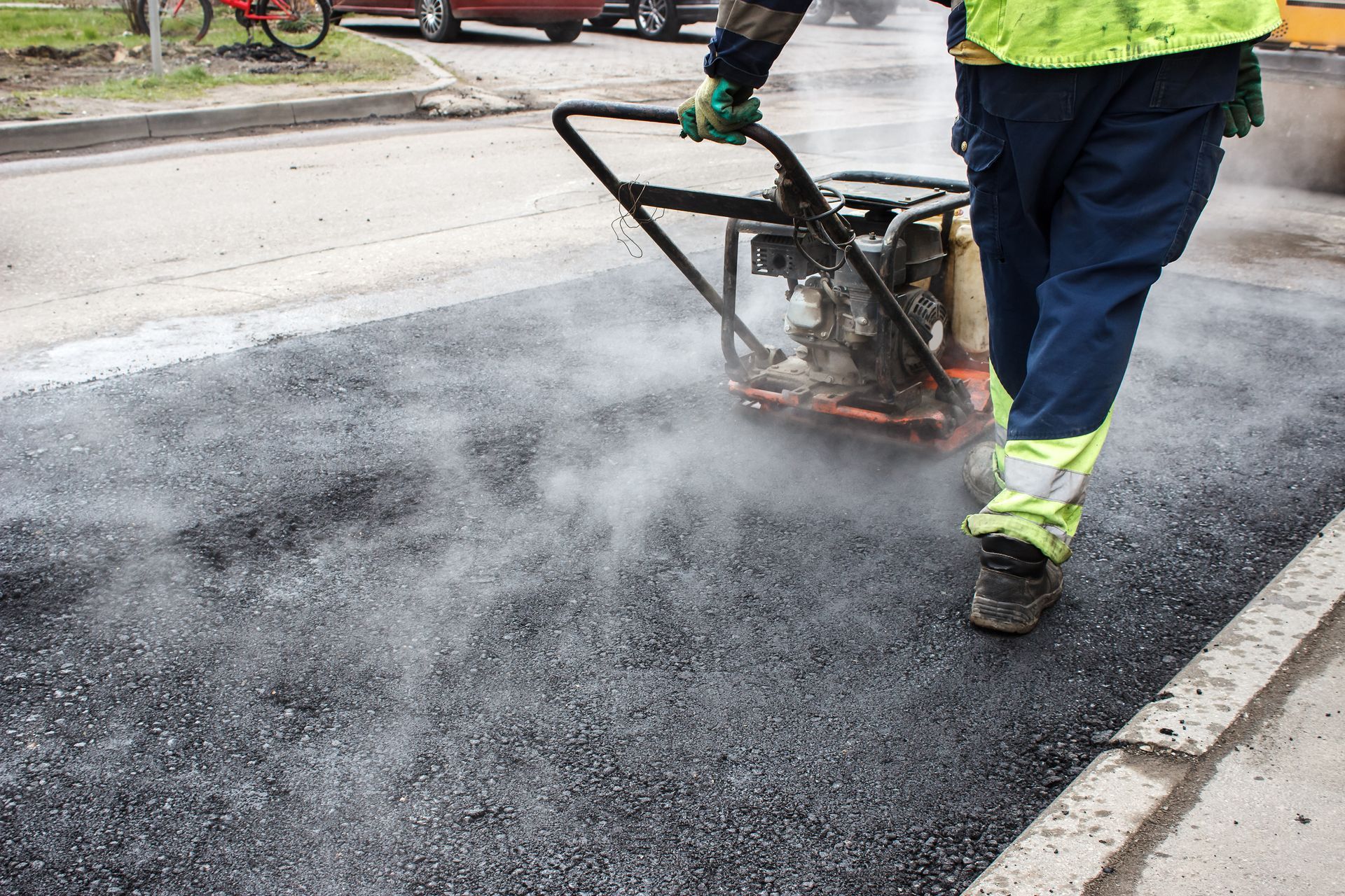 Worker operating a vibrating plate compactor on freshly paved asphalt, with steam or dust rising nearby Worker operating a vibrating plate compactor on freshly paved asphalt, with steam or dust rising nearby