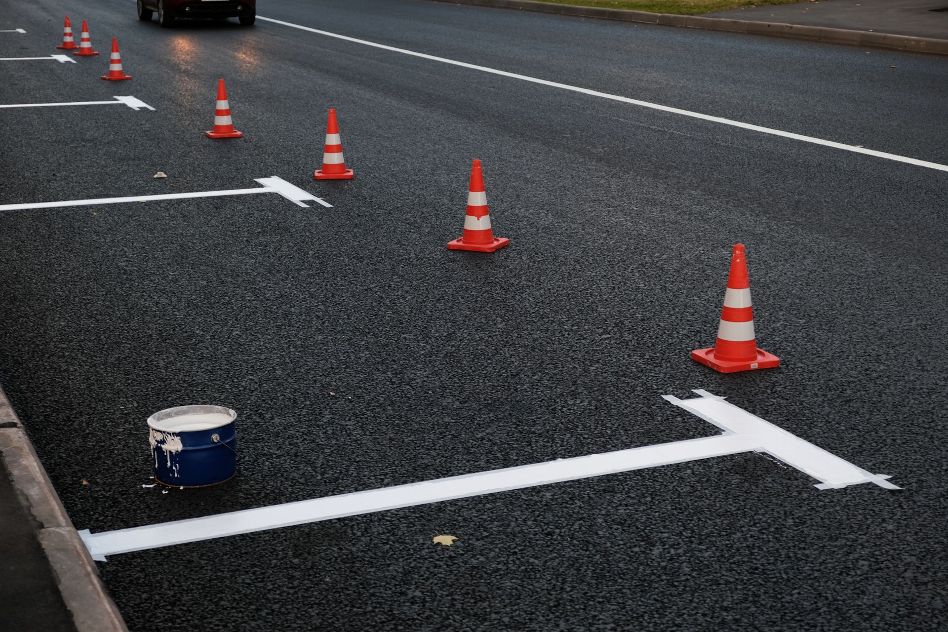 Traffic cones marking white lane lines on wet asphalt, with a small paint can in the foreground.