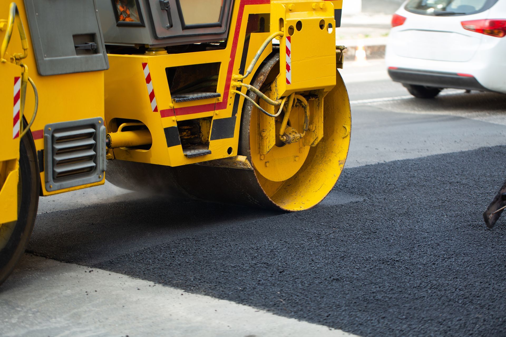 Yellow road roller paving dark asphalt beside a white car on a street Yellow road roller paving dark asphalt beside a white car on a street