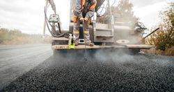 Road paving machine laying fresh black asphalt on a road, with dust and construction equipment visible