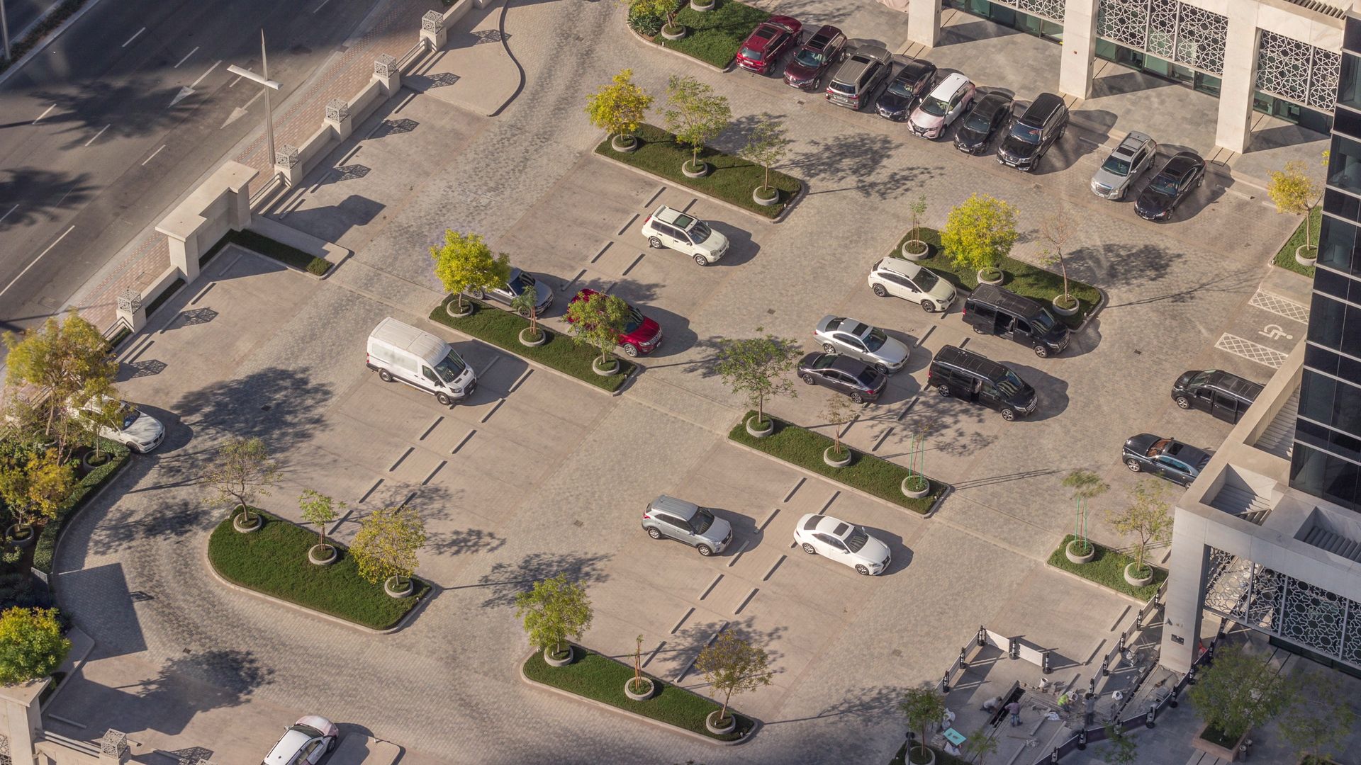 Aerial view of a parking lot with parked cars, trees, and marked spaces beside a road and building. Aerial view of a parking lot with parked cars, trees, and marked spaces beside a road and building.