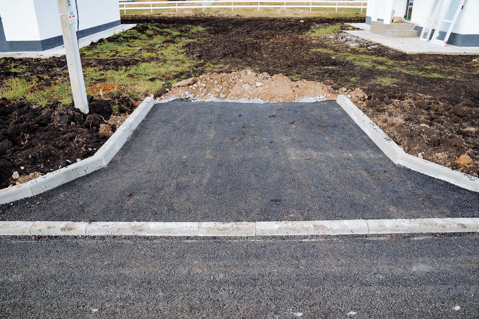 Freshly paved black asphalt ramp between curbs leading to a dirt path and building entrance