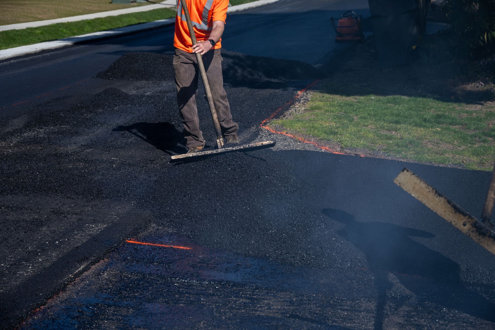 Worker in orange vest smoothing fresh black asphalt on a road.