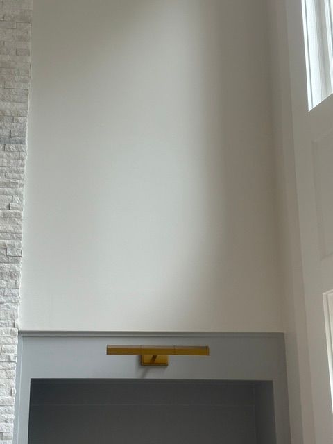 A tall white wall above a gray fireplace, with a gold light fixture mounted on top.