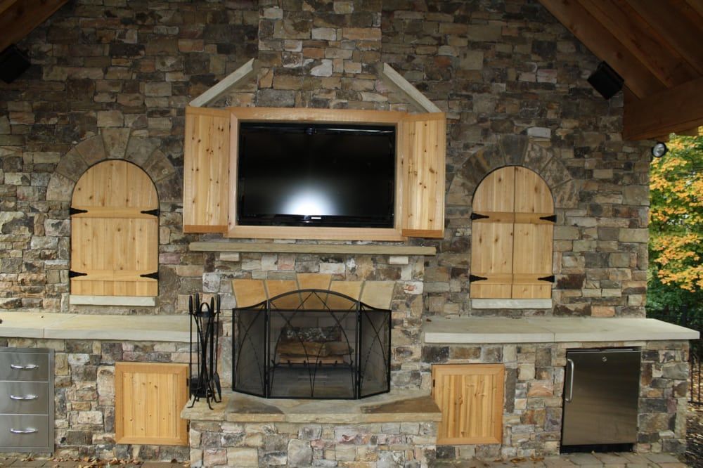 Outdoor stone fireplace with TV, shutters, and cabinetry.
