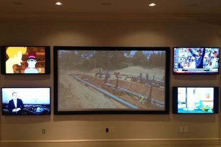 Wall with six TVs displaying various programs. A large black-framed TV is centered.