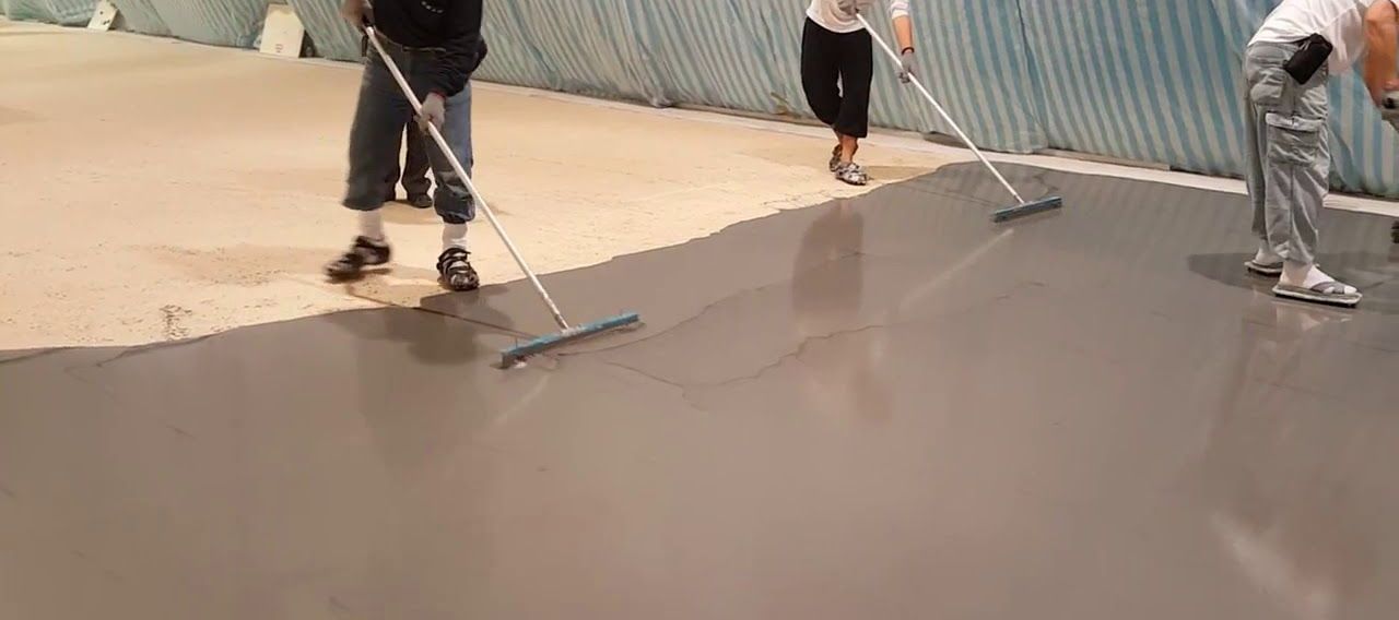 Workers smoothing wet concrete with long-handled tools on a large floor slab.