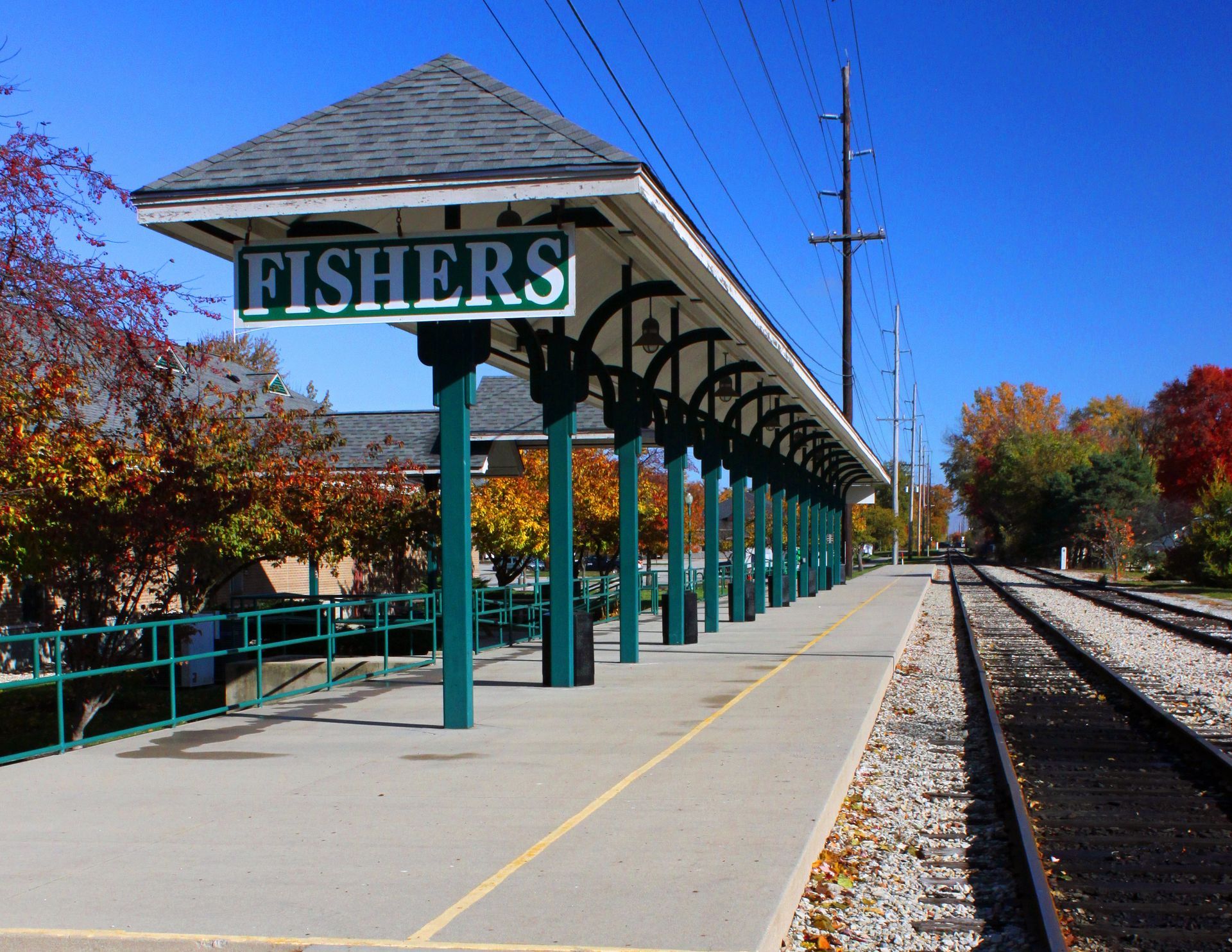 Fishers train station platform with green sign, covered walkway, and railroad tracks on a sunny day