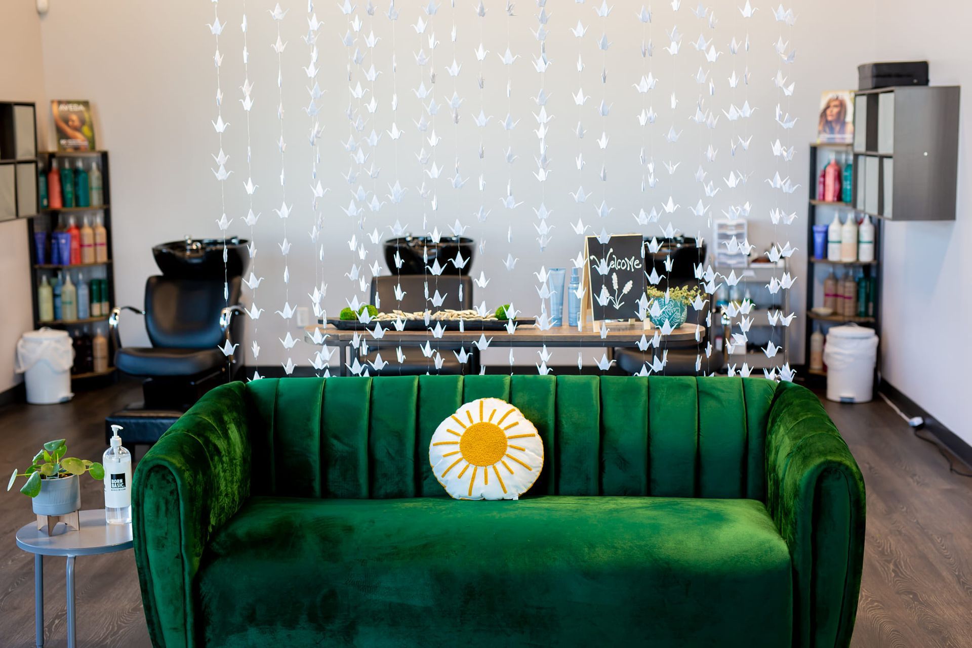 A green velvet sofa with a sun-shaped pillow sits in a hair salon with white hanging paper cranes in the background.