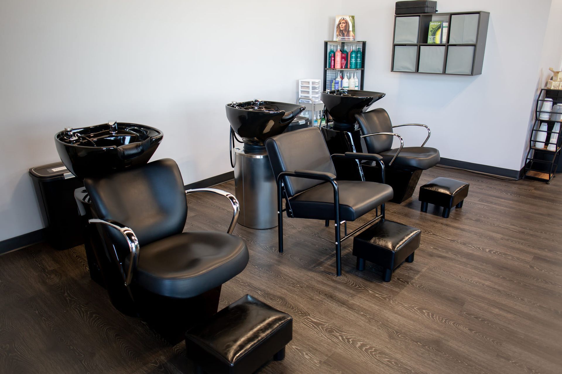 Three black salon shampoo stations with chairs and footrests arranged in a row in a clean, minimalist salon space.