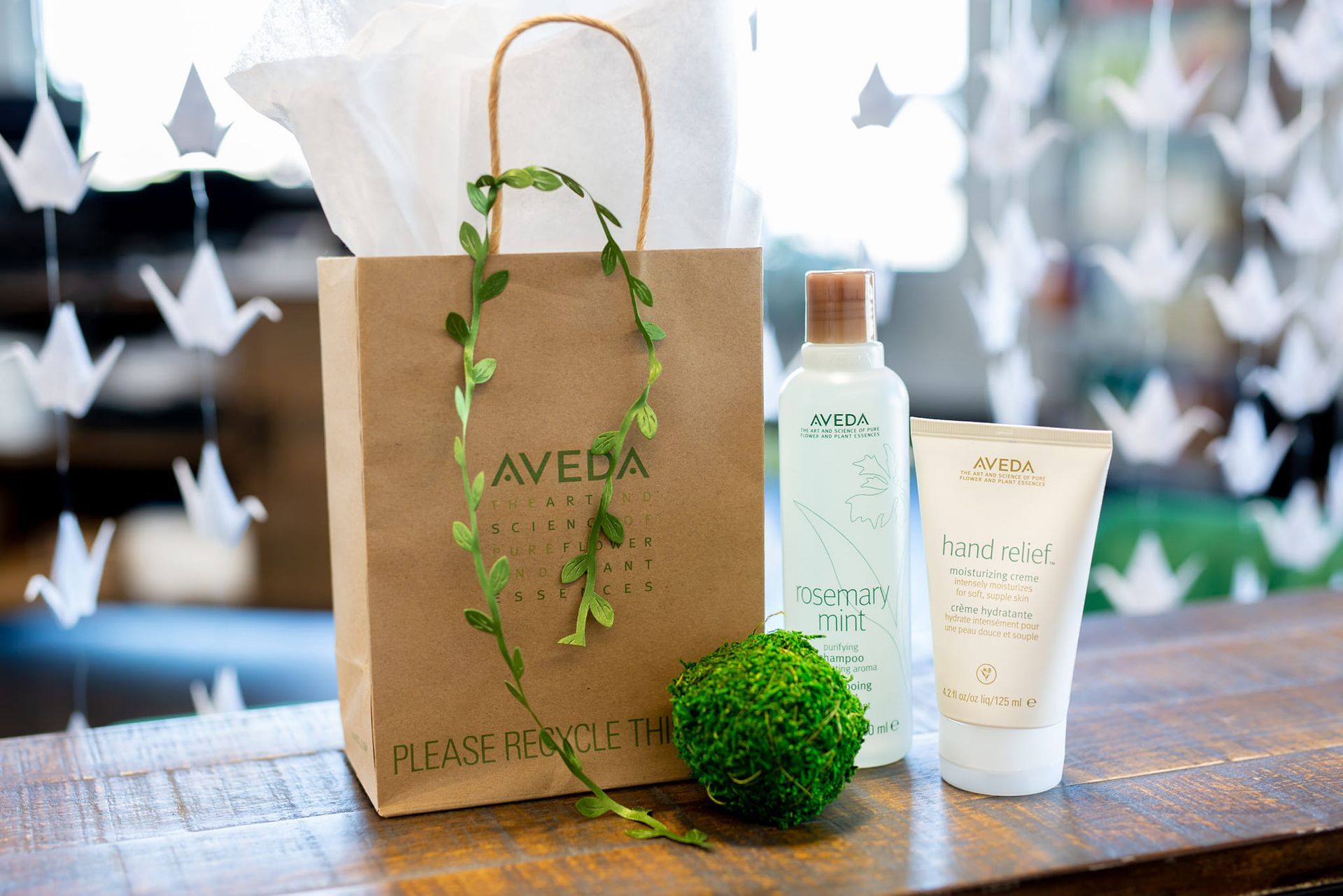 A brown Aveda paper bag, a bottle of Aveda hair product, and a tube of hand cream on a wooden counter with origami birds.
