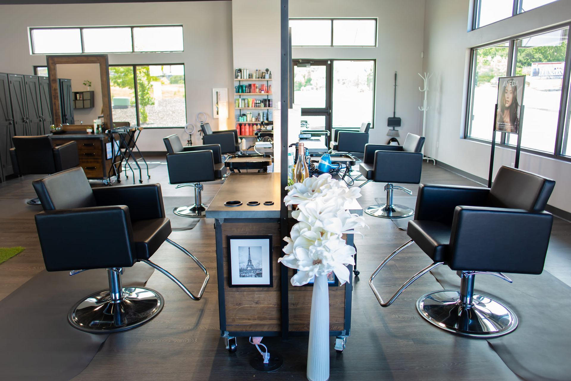 Interior of a brightly lit hair salon featuring several black styling chairs facing mirrors in a modern, open space.