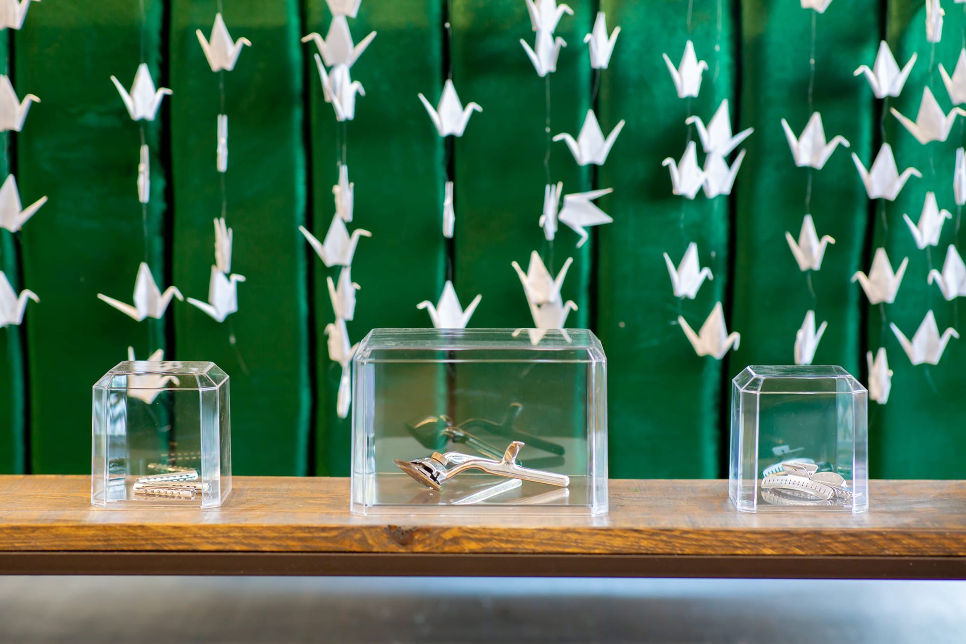 Three clear display cases on a wooden shelf in front of a green backdrop decorated with hanging white paper cranes.
