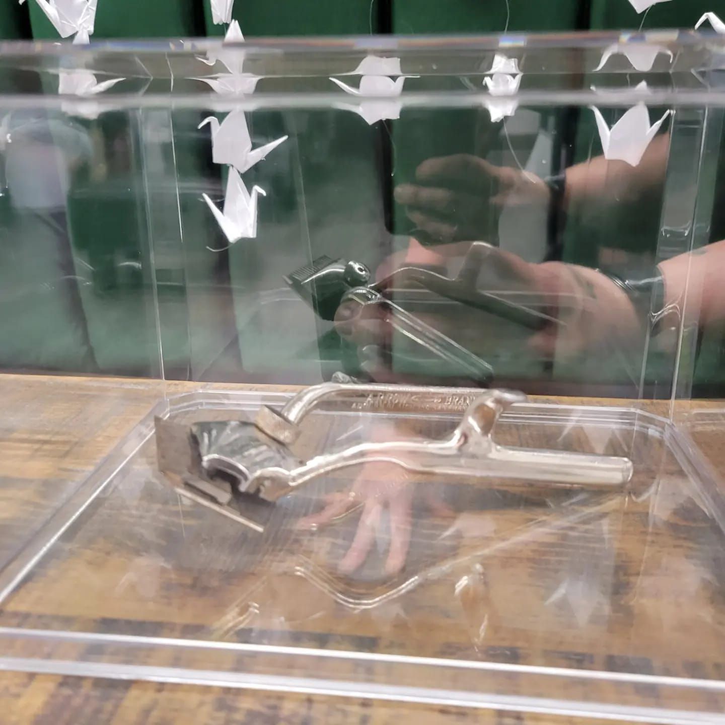 A silver metal garlic press rests inside a clear acrylic display case near white paper cranes.
