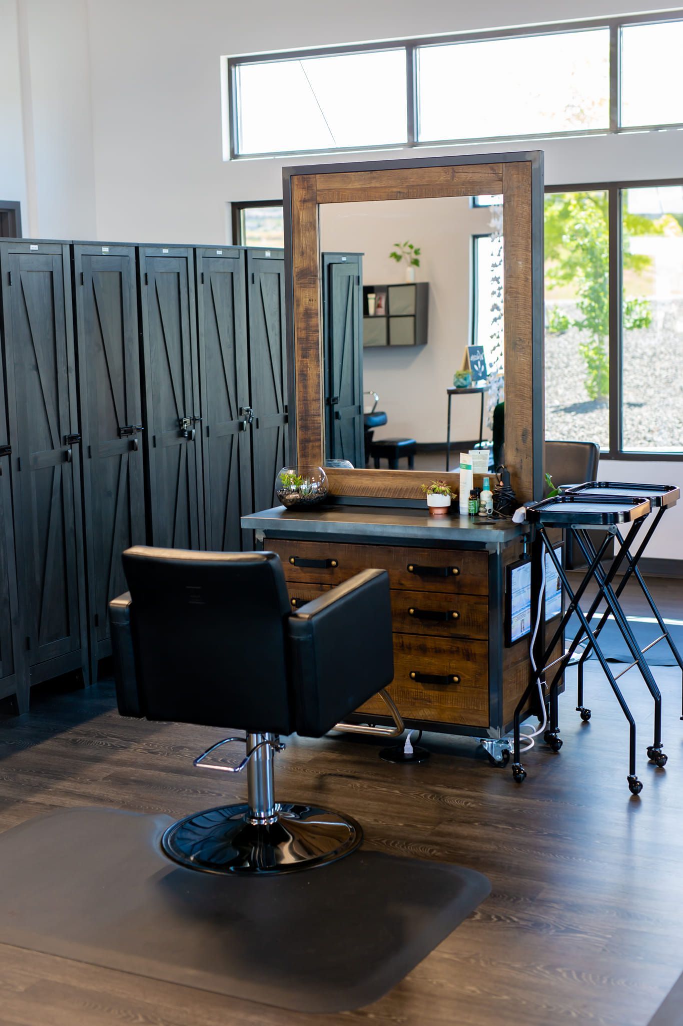 A professional salon station featuring a black chair, a rustic wooden mirror, drawers, and a row of dark metal lockers.